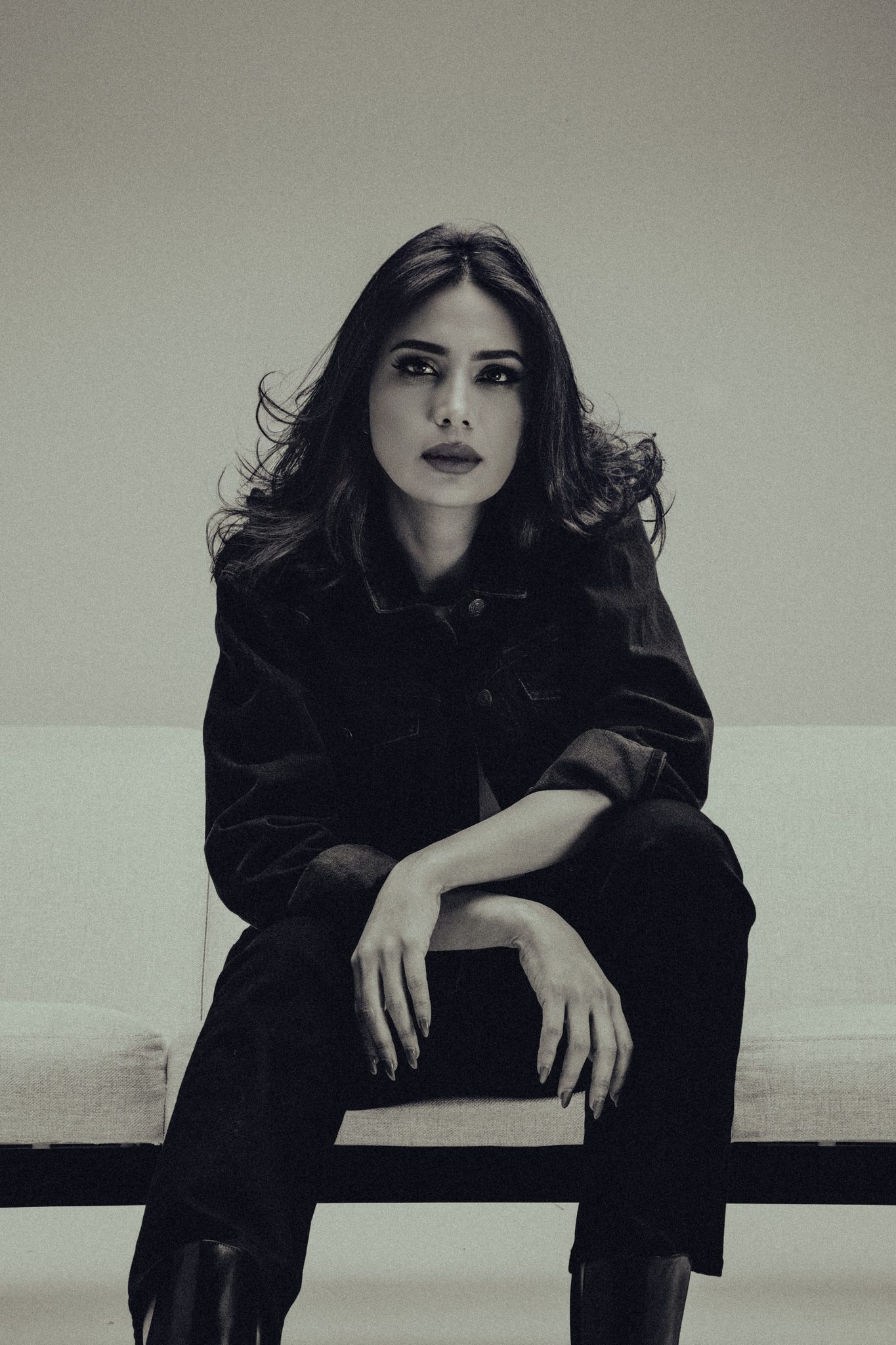 Black-and-white fashion editorial of a woman in an oversized black jacket seated with hands clasped on a minimalist bench